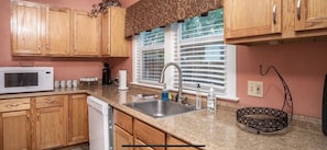A kitchen with wooden cabinets, a microwave, a sink, and a window with blinds.