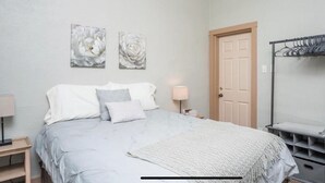 A bedroom with a bed, two wall-mounted paintings, a door, a bedside table with a lamp, and a metal drying rack. - Spacious R&R University Retreat  (Pocatello)