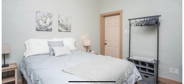 A bedroom with a bed, two wall-mounted paintings, a door, a bedside table with a lamp, and a metal drying rack.