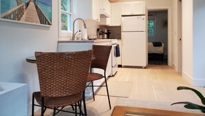 A modern kitchen with white cabinets, a built-in oven, and a refrigerator. There are wicker chairs and a dining table. - One Bedroom Hamptons Waterfront Cottage (Hampton Bays)