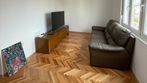 Una sala de estar con piso de madera en espiga, un sofá de cuero marrón, un televisor sobre un mueble de madera y una lámpara colgante.