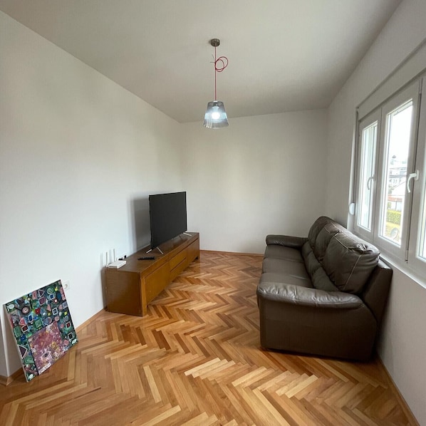 A living room with a herringbone wood floor, a brown leather sofa, a TV on a wooden stand, and a pendant light.