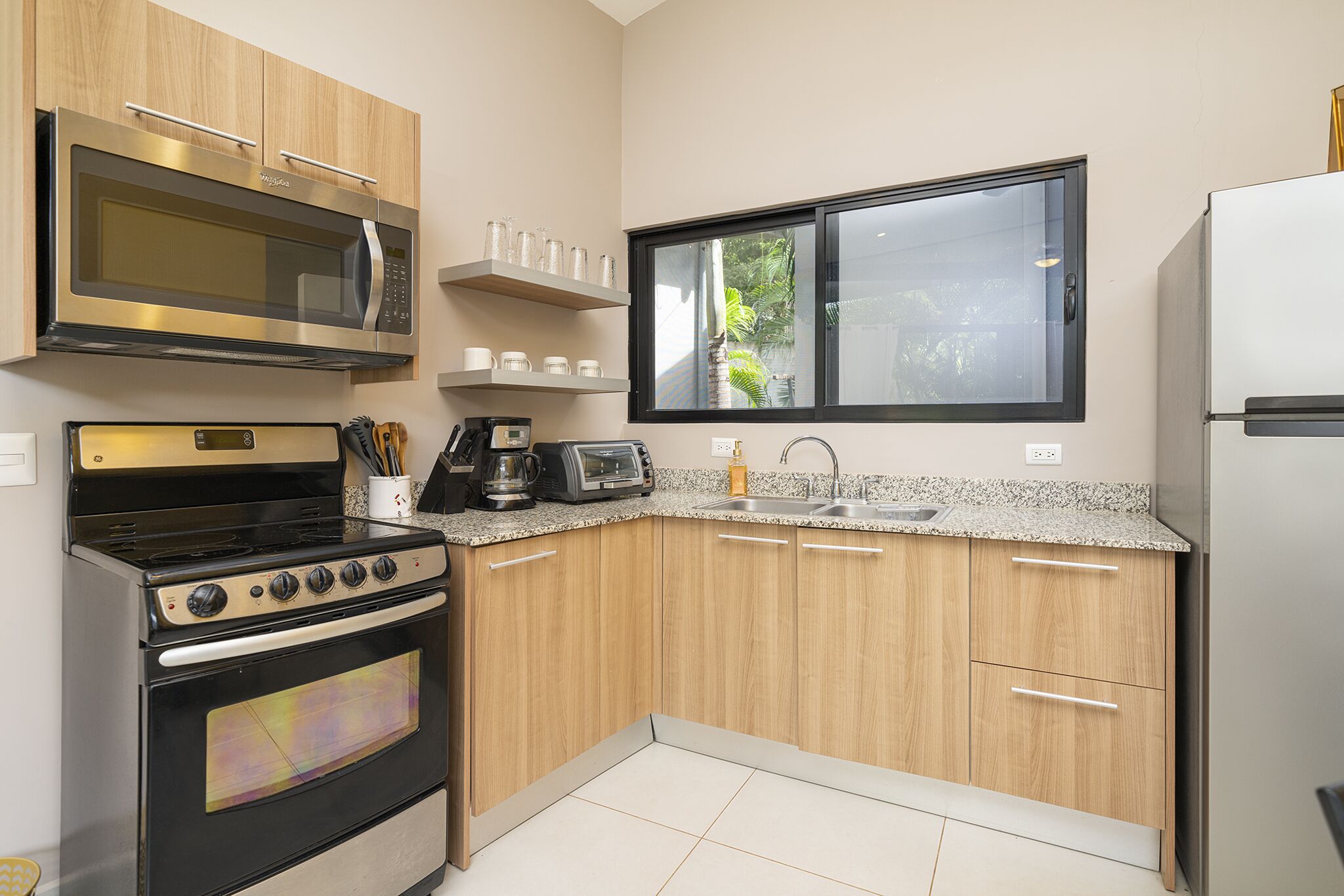 A compact kitchen with a microwave, stove, and oven, wooden cabinets, a granite countertop, a window, and a refrigerator.