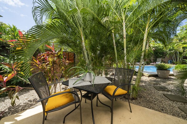 An outdoor patio area with a table and chairs, surrounded by lush greenery and a pool.