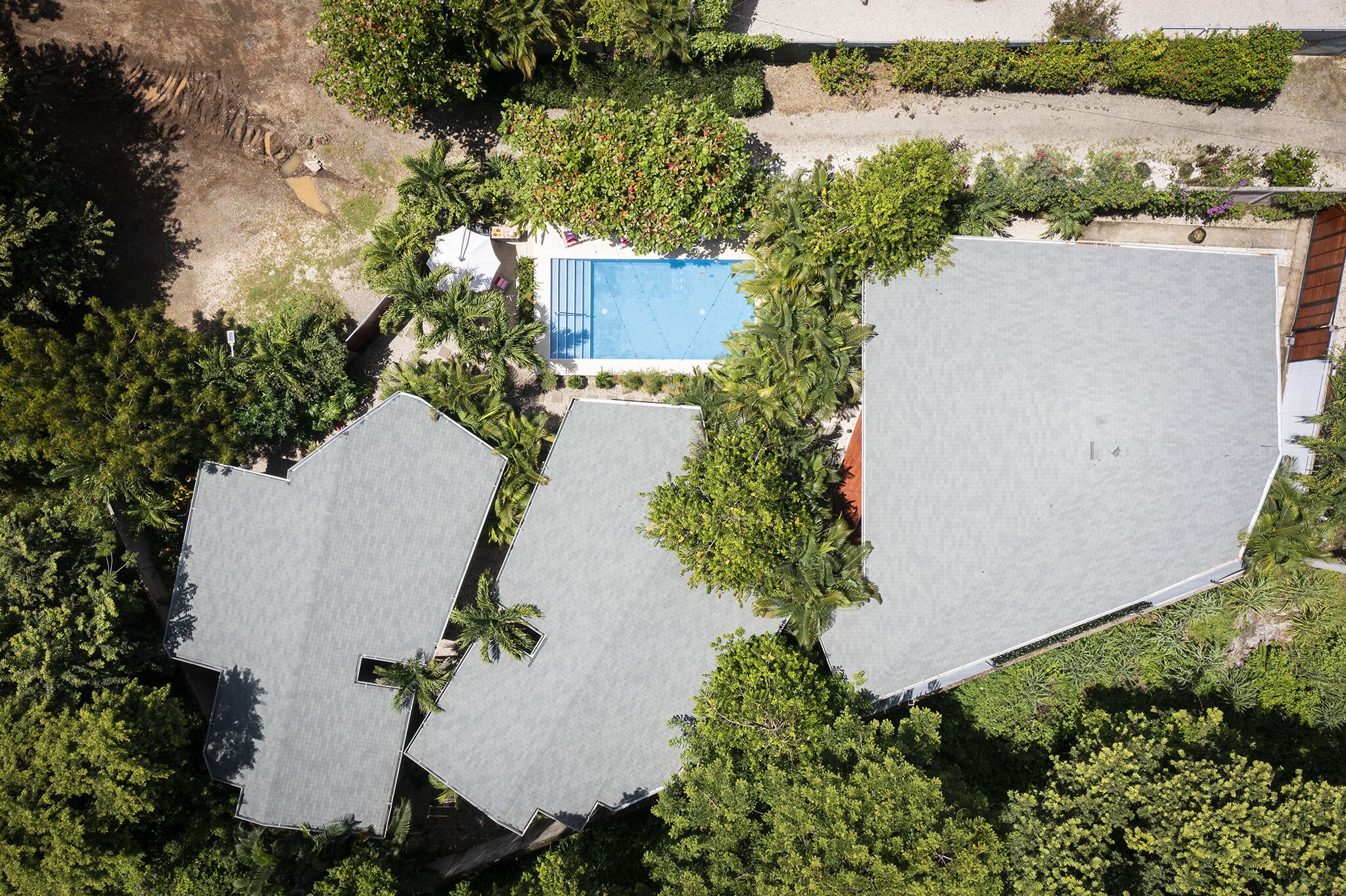 Ovenudblik over en boligejendom med swimmingpool omgivet af frodigt grønt, flere tagområder og en lille have.