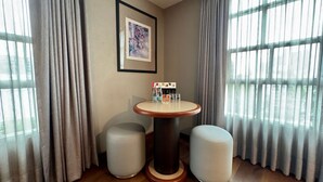 Quarto com uma mesa redonda, dois pufes, um quadro emoldurado e uma janela com cortinas.