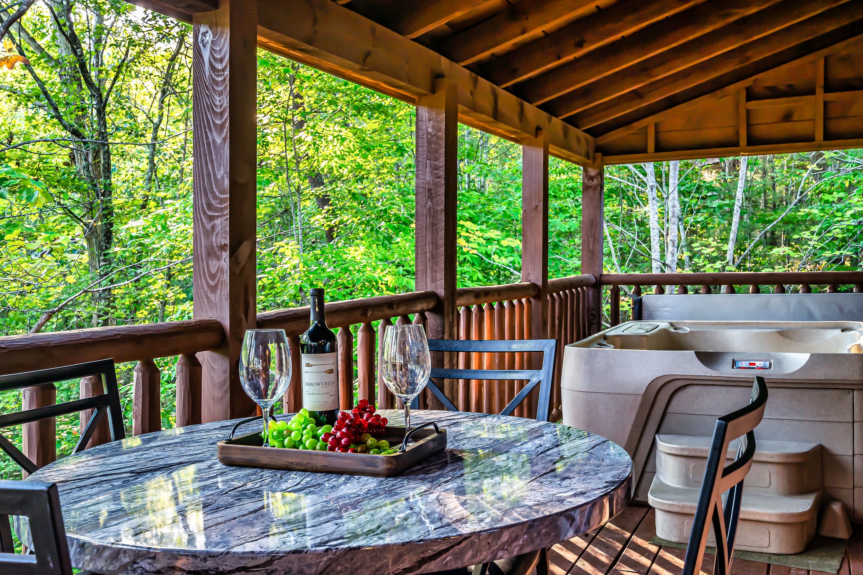 Un espacio al aire libre techado con mesa lista para comer, jacuzzi y vista a un bosque.