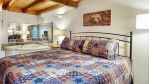 Ein Schlafzimmer mit einem großen Bett, einer Holzbalkendecke, einem Gemälde an der Wand und einem Badezimmer mit Spiegel und Waschbecken.