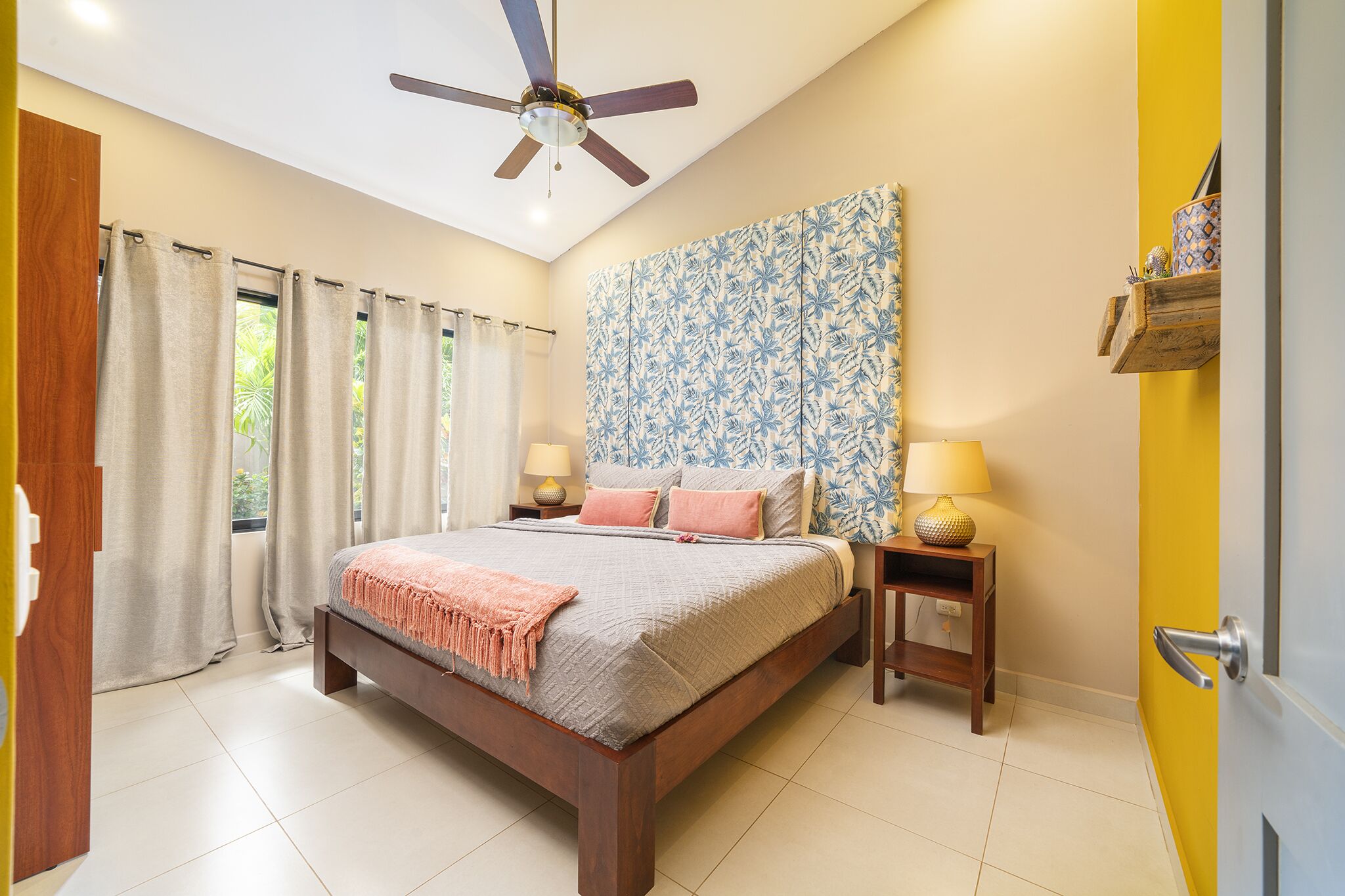 A bedroom with a bed, a ceiling fan, a nightstand with a lamp, and a window with curtains.