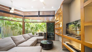A modern living room with a large sectional sofa, a flat-screen TV on a wooden shelf, and a view of lush greenery through large windows. - Total Stellar Entire Property 3 Villas Luxurious Oasis Getaway (Playa Langosta, Tamarindo)