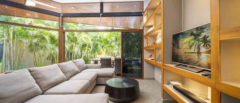 A modern living room with a large sectional sofa, a flat-screen TV on a wooden shelf, and a view of lush greenery through large windows.