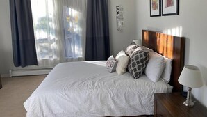 Une chambre à coucher bien rangée, avec un grand lit, une table de chevet, une lampe et une fenêtre avec des rideaux.