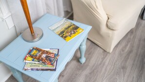 Ein heller blauer Tisch mit Zeitschriften und einer Lampe, ein beiger Sessel mit einem gemusterten Kissen und ein Holzboden.