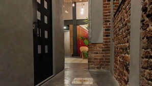 Un pasillo estrecho con una pared de ladrillo a la derecha, una puerta negra a la izquierda y una puerta roja al final.