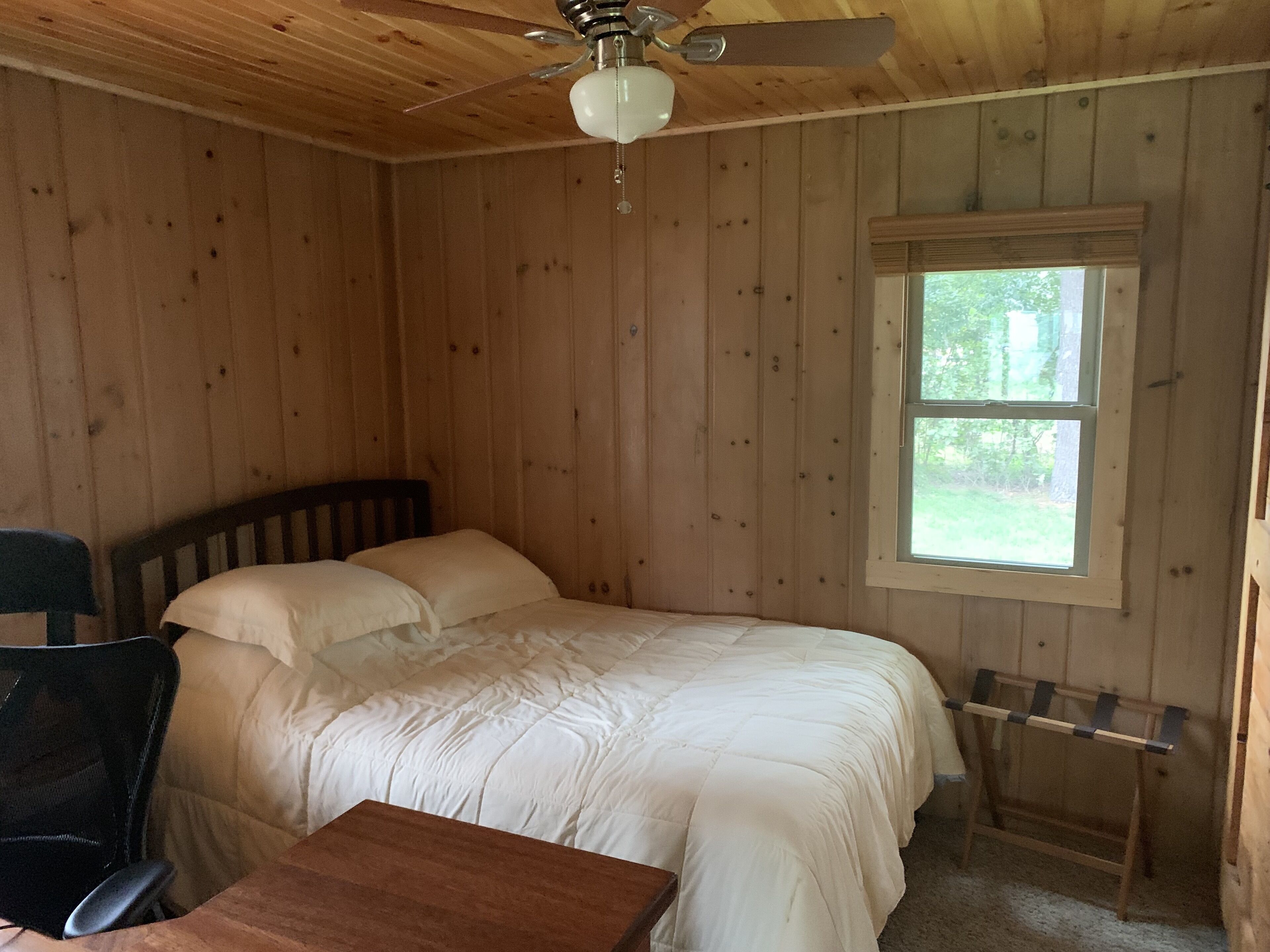 A wooden cabin bedroom with a bed, a desk, a chair, a ceiling fan, and a window.