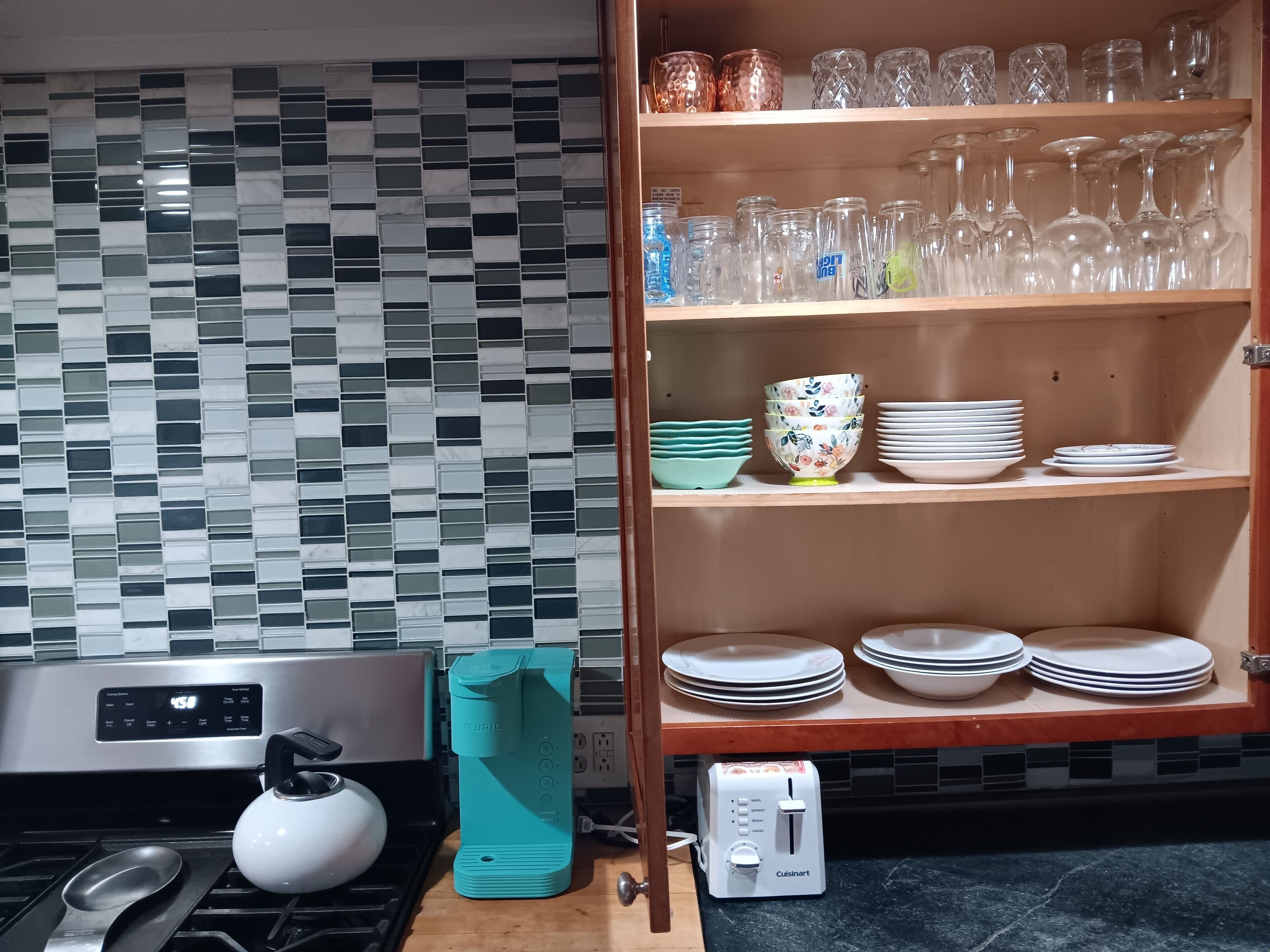 A kitchen with a tiled backsplash, a stove, a kettle, a toaster, and a shelf with glassware and dishes.
