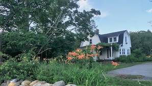 Une maison blanche avec un toit foncé, entourée d’un mur de pierre et d’un jardin où poussent des fleurs oranges.