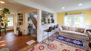 Un salon chaleureux avec un canapé, une table basse et un tapis à motifs. Un escalier mène à un autre niveau, et une bibliothèque est remplie de livres et d’objets décoratifs.