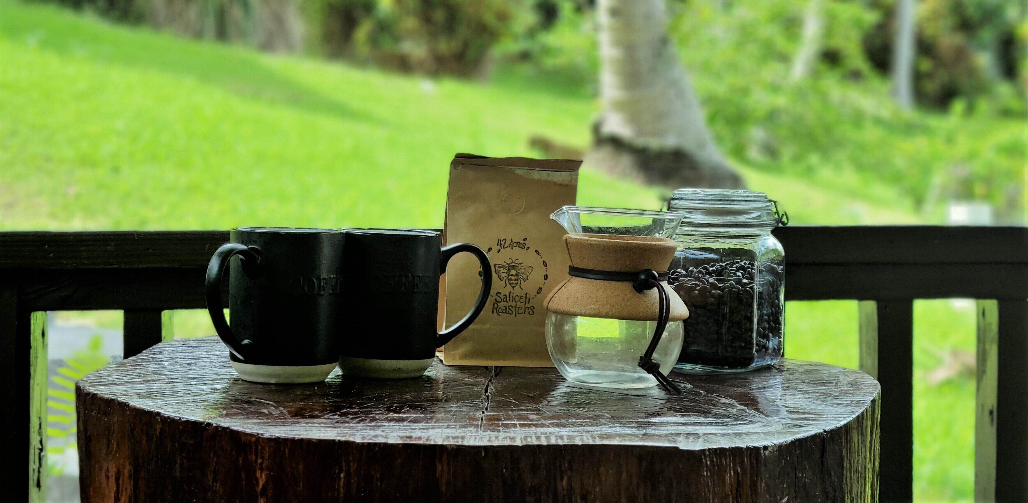 A rustic wooden table with two black mugs, a paper bag labeled "Salisch Roasters," and a glass jar with coffee beans.