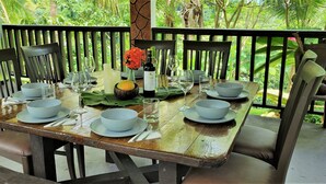 A dining table set with plates, bowls, wine glasses, and a bottle, surrounded by chairs, with a view of lush greenery. - Tropical Rainforest Retreat (Naguabo)