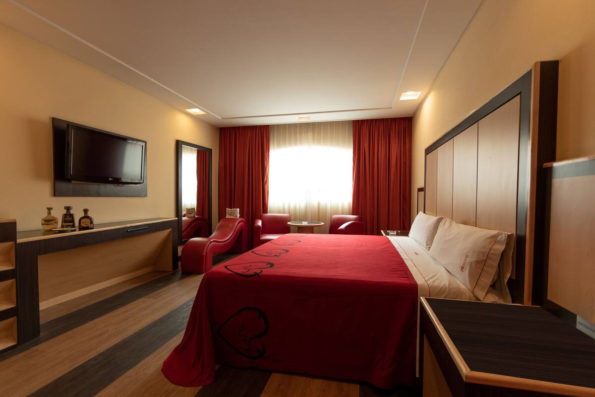 Une chambre d’hôtel avec un lit, un téléviseur fixé au mur, un bureau avec des bouteilles et une fenêtre avec des rideaux.
