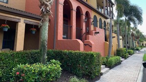 Una hilera de edificios con ventanas y balcones arqueados, rodeada de setos cuidados y palmeras.