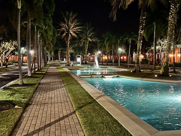 A nighttime pool area with a paved walkway, palm trees, and a fountain.