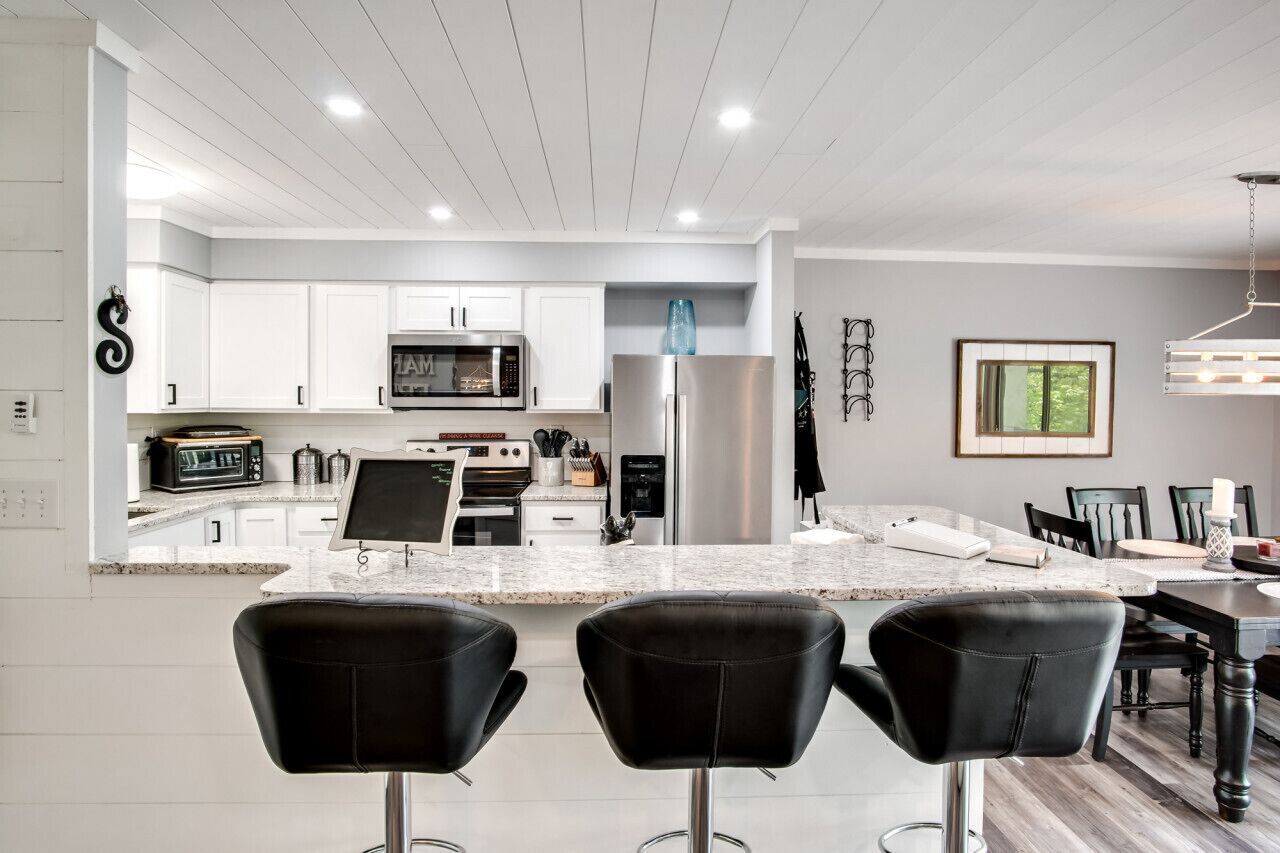 A modern kitchen with white cabinetry, a stainless steel refrigerator, a microwave, and a built-in oven. There are three black bar stools in front of the kitchen island.