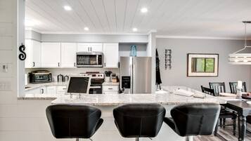 A modern kitchen with white cabinetry, a stainless steel refrigerator, a microwave, and a built-in oven. There are three black bar stools in front of the kitchen island.
