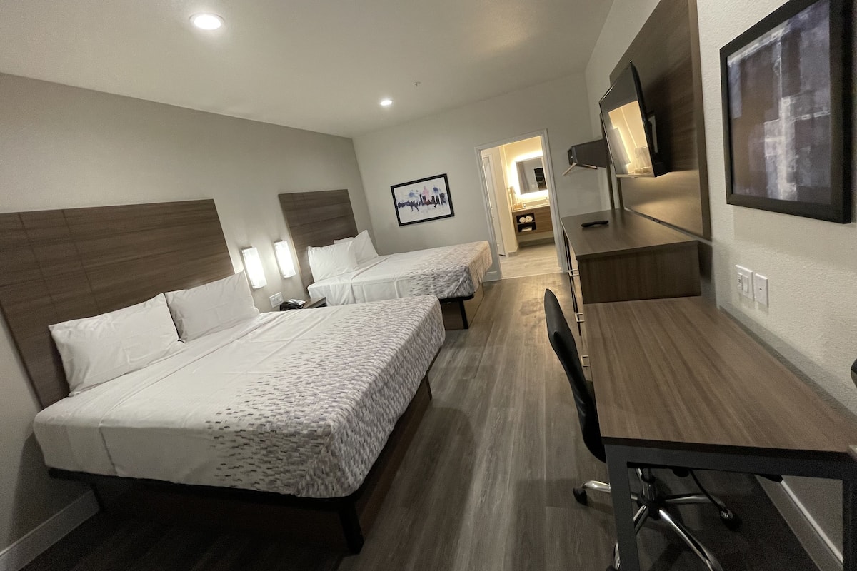 Ett hotellrum med två sängar, ett skrivbord, en stol, en tv och ett badrum som syns i bakgrunden.