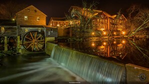 Un molino histórico con una rueda hidráulica, edificios de madera y un restaurante llamado "The Old Mill Restaurant" junto a un río, de noche.