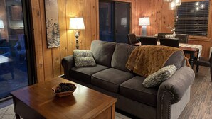 Un salon chaleureux avec un mur lambrissé en bois, un canapé gris agrémenté d’un plaid marron, deux lampes et, au fond, un coin repas.