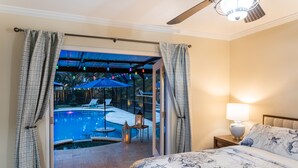 Un dormitori amb un llit, una tauleta de nit, una làmpada i vista a una zona de piscina exterior.