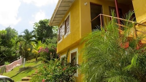 A two-story yellow building with a red roof, surrounded by lush greenery and palm trees. - La Familia Guesthouse (Long Bay)