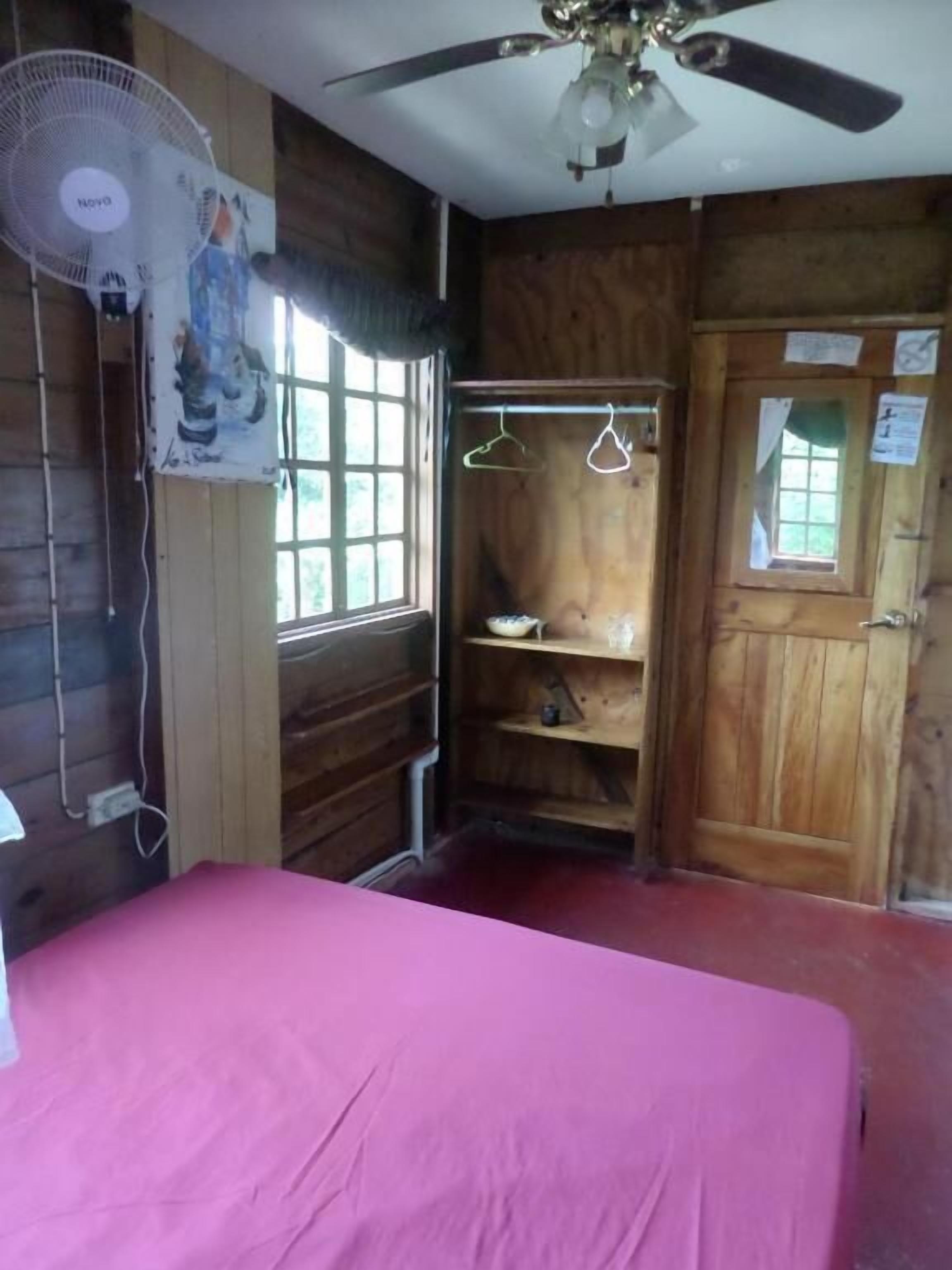 Quarto com paredes de madeira, uma cama com colcha rosa, um ventilador de teto, uma janela com vista e uma porta de madeira com uma pequena janela.
