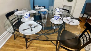 Une table ronde en verre, dressée pour six personnes, avec des assiettes blanches, des tasses à motifs floraux bleus et des couverts.