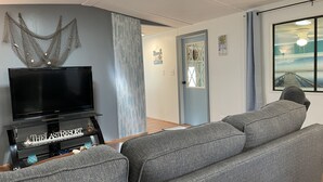 Un soggiorno con una TV a schermo piatto su un mobile, divani grigi imbottiti, un pannello decorativo e vista sulla spiaggia attraverso una finestra.