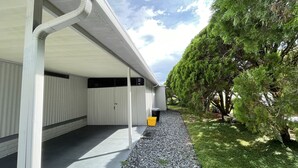 Un'area esterna coperta con pavimento in cemento, un cestino e un vialetto sterrato che conduce a un edificio con facciata bianca e un grande albero nelle vicinanze.