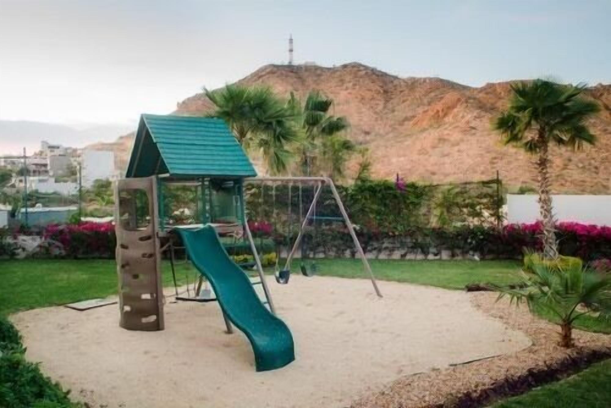 A playground with a slide and swings, surrounded by greenery and a hill in the background.