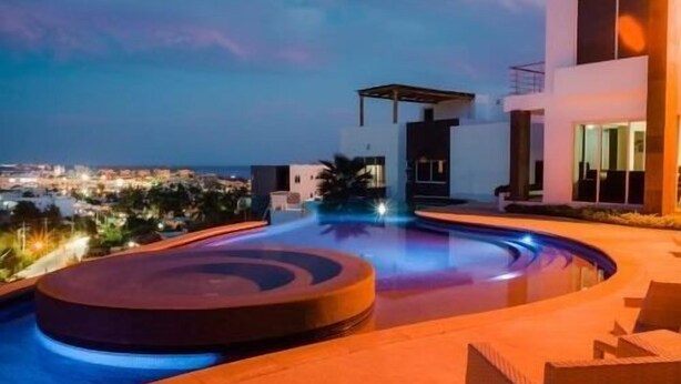 Un área de piscina exterior moderna con diseño circular, un espacio de estar cubierto y vistas a la ciudad al anochecer.