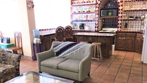 A living room with a sofa, a glass-top coffee table, and a kitchen area with a tiled backsplash and wooden cabinets.