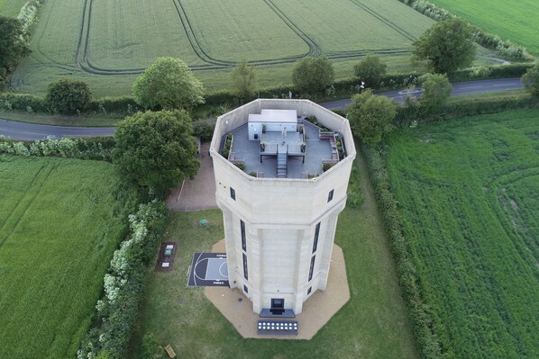 Una torre cilíndrica con techo plano, rodeada de campos verdes y una carretera.