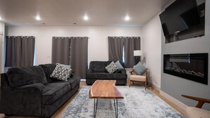 Una sala de estar moderna con dos sofás oscuros, una mesa de centro de madera, un sillón pequeño y un televisor de pantalla plana montado en la pared.