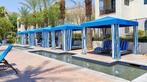 A pool area with blue umbrellas and lounge chairs, surrounded by palm trees and a building with balconies. - Relaxing Desert Villa in Resort Style property (La Quinta)