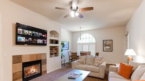 A living room with a fireplace, a flat-screen TV mounted on the wall, a ceiling fan, and a large window with blinds. - Relaxing Desert Villa in Resort Style property (La Quinta)