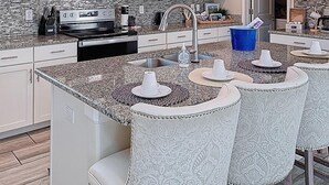 Cozinha com armários brancos, micro-ondas, fogão e bancada de granito. Há duas luminárias pendentes e um balde azul.