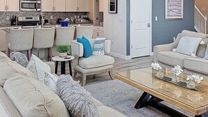 Uma sala de estar moderna com um sofá, poltronas, uma mesa de centro e uma cozinha ao fundo.