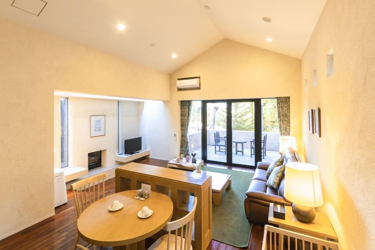 A spacious living area with a wooden dining table, a sofa, and a TV. There is a sliding glass door leading to an outdoor patio.