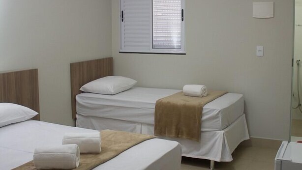 A hotel room with two beds, a window with blinds, an air conditioning unit, and a small white cabinet. - Hotel Alphaville (Ouvidor)