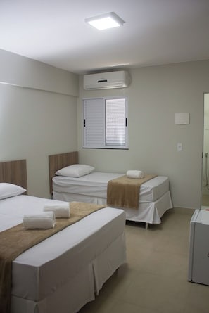 Ett hotellrum med två sängar, ett fönster med persienner, en luftkonditioneringsanläggning och ett litet kylskåp.